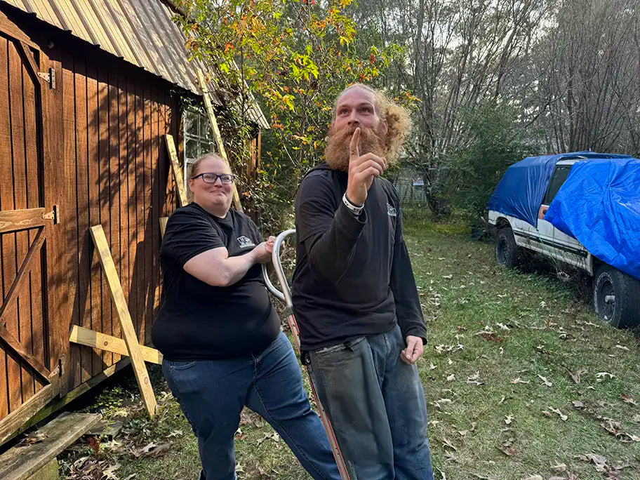 Two people standing outdoors next to a wooden shed with trees and a covered vehicle in the background.